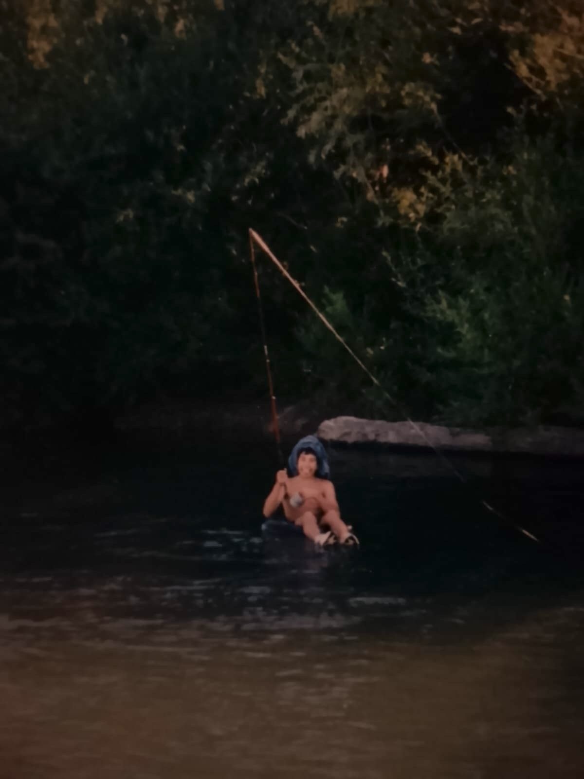 A child floating on an inner tube in a dark river, holding a long fishing rod at dusk