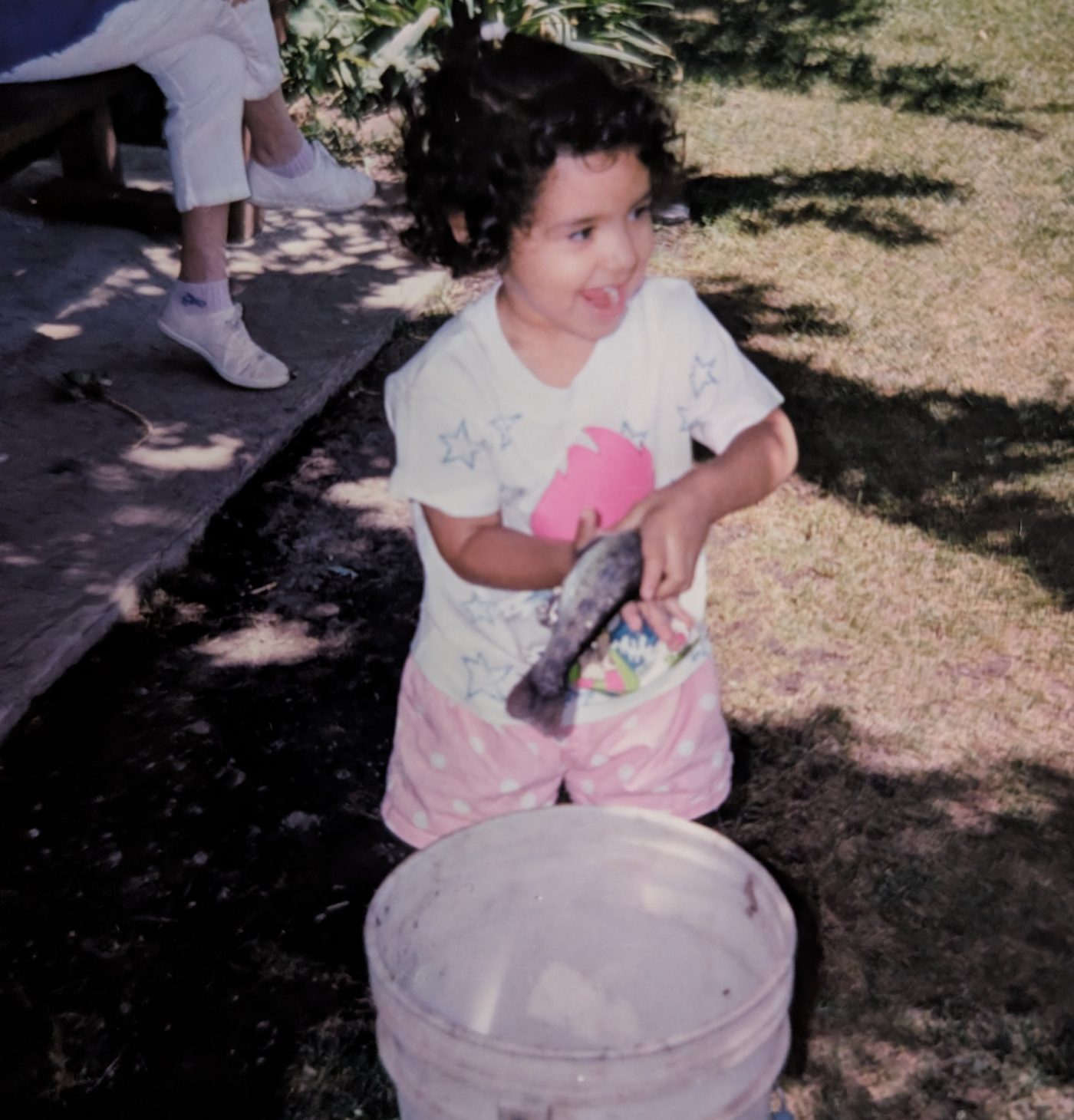 A laughing toddler with curly hair holding a fish over a bucket in a sunny garden