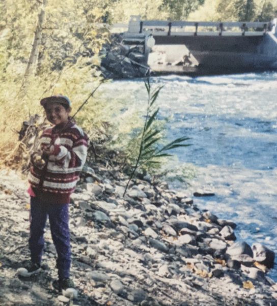 A smiling child in a striped sweater and cap holding a fishing rod on a rocky riverbank