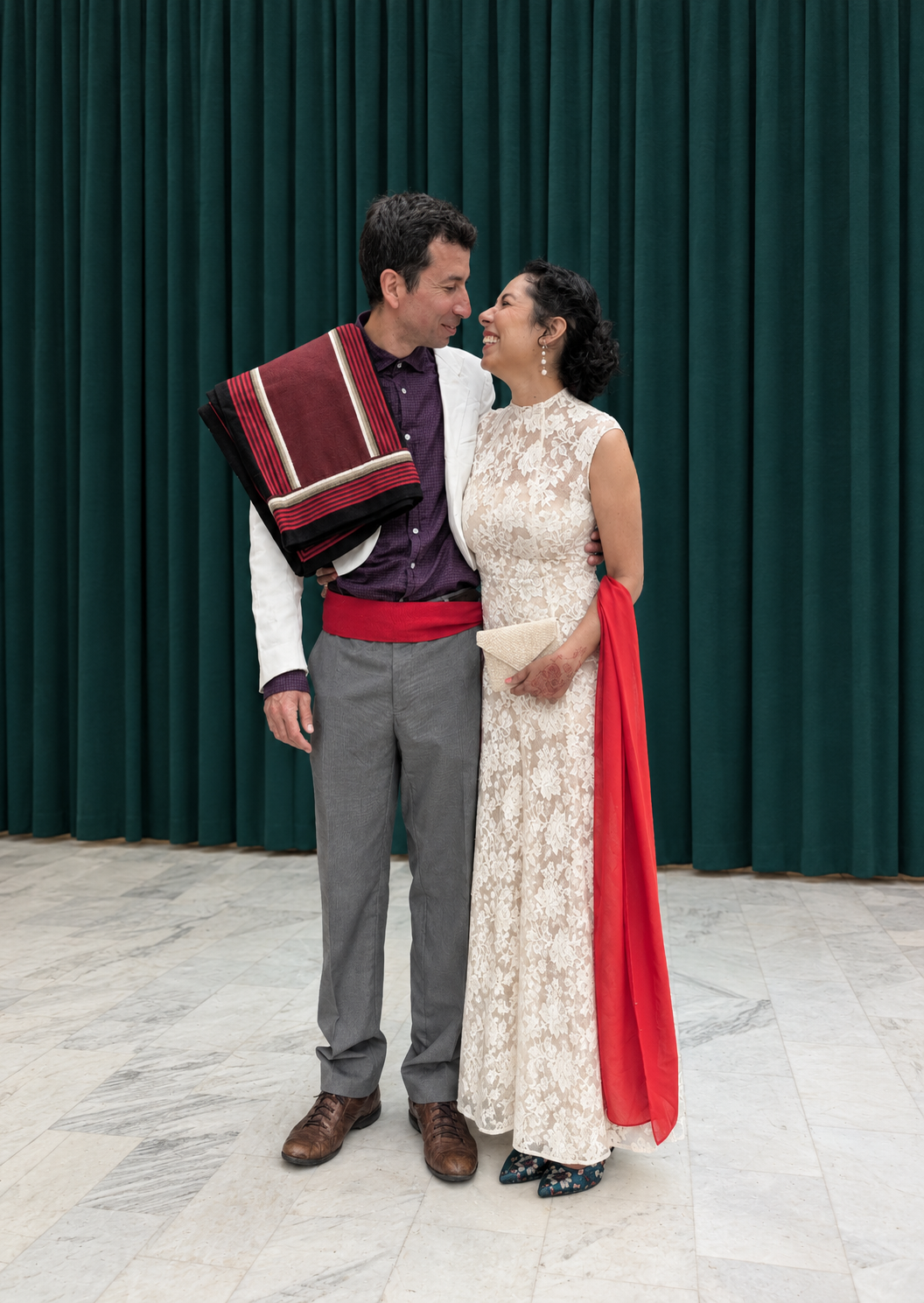 Sabah in a white lace gown and red shawl and Jose Manuel in a white jacket and striped shawl, smiling at each other in front of a green velvet curtain