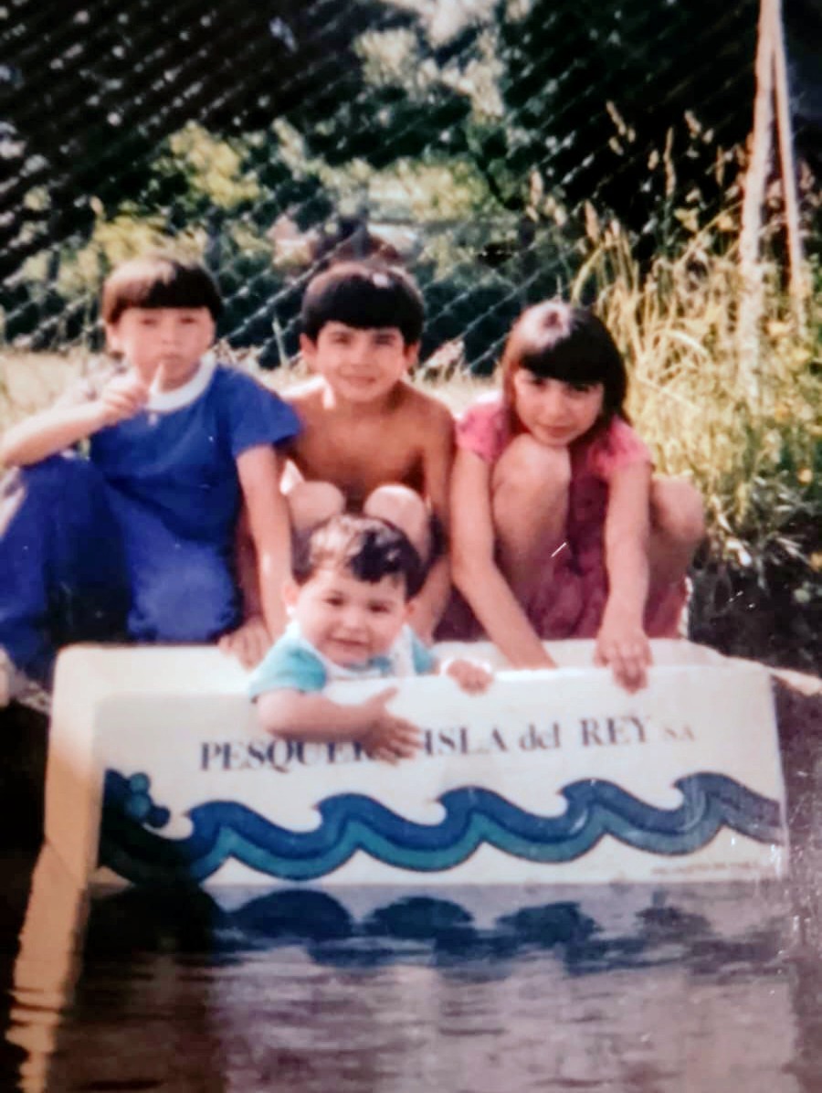 Four young children by the water, the smallest sitting in a white Pesquera Isla del Rey box
