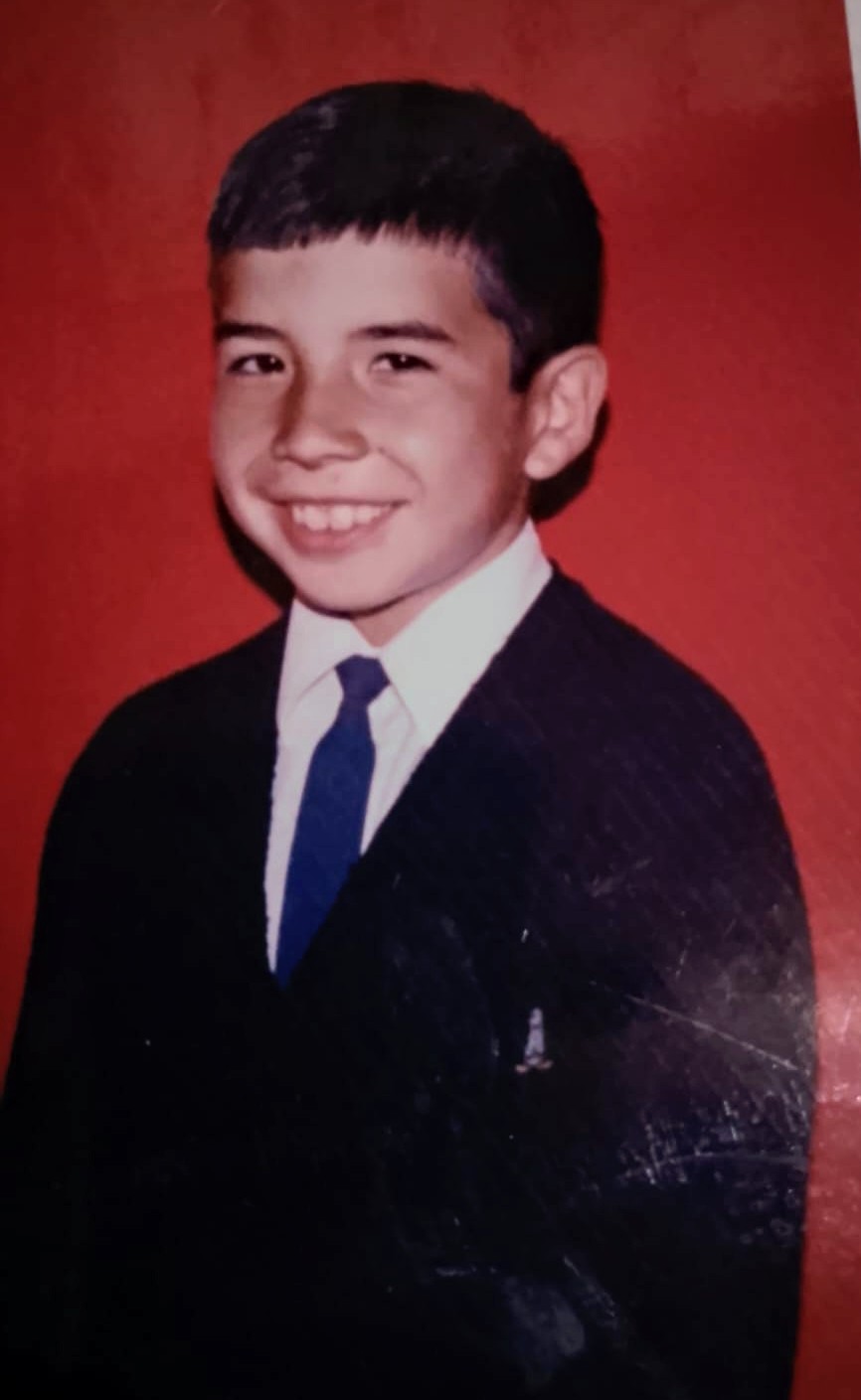 A school portrait of a smiling young Jose Manuel in a navy blazer and blue tie against a red backdrop