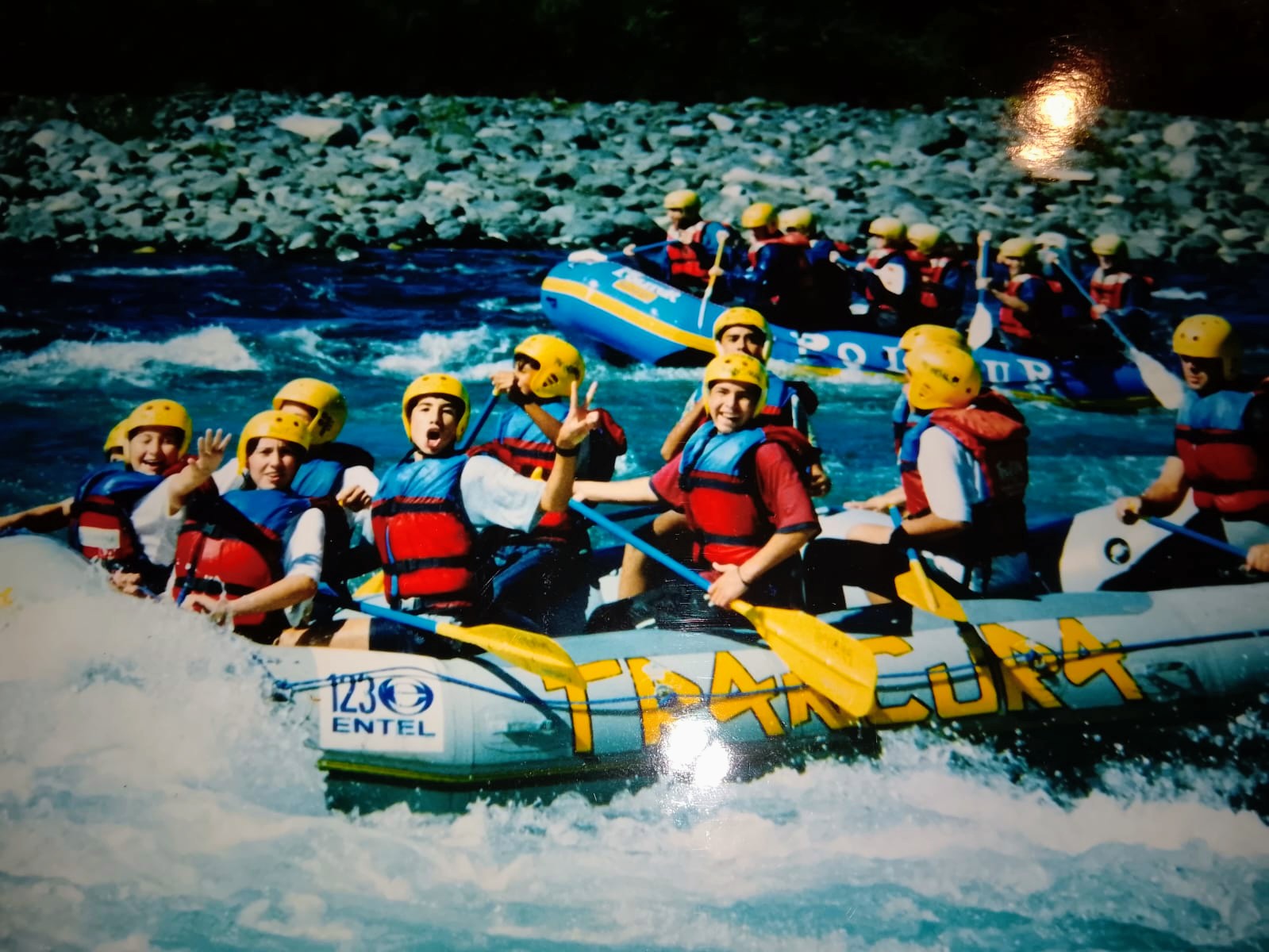 A raft of teenagers in yellow helmets and life jackets cheering through whitewater