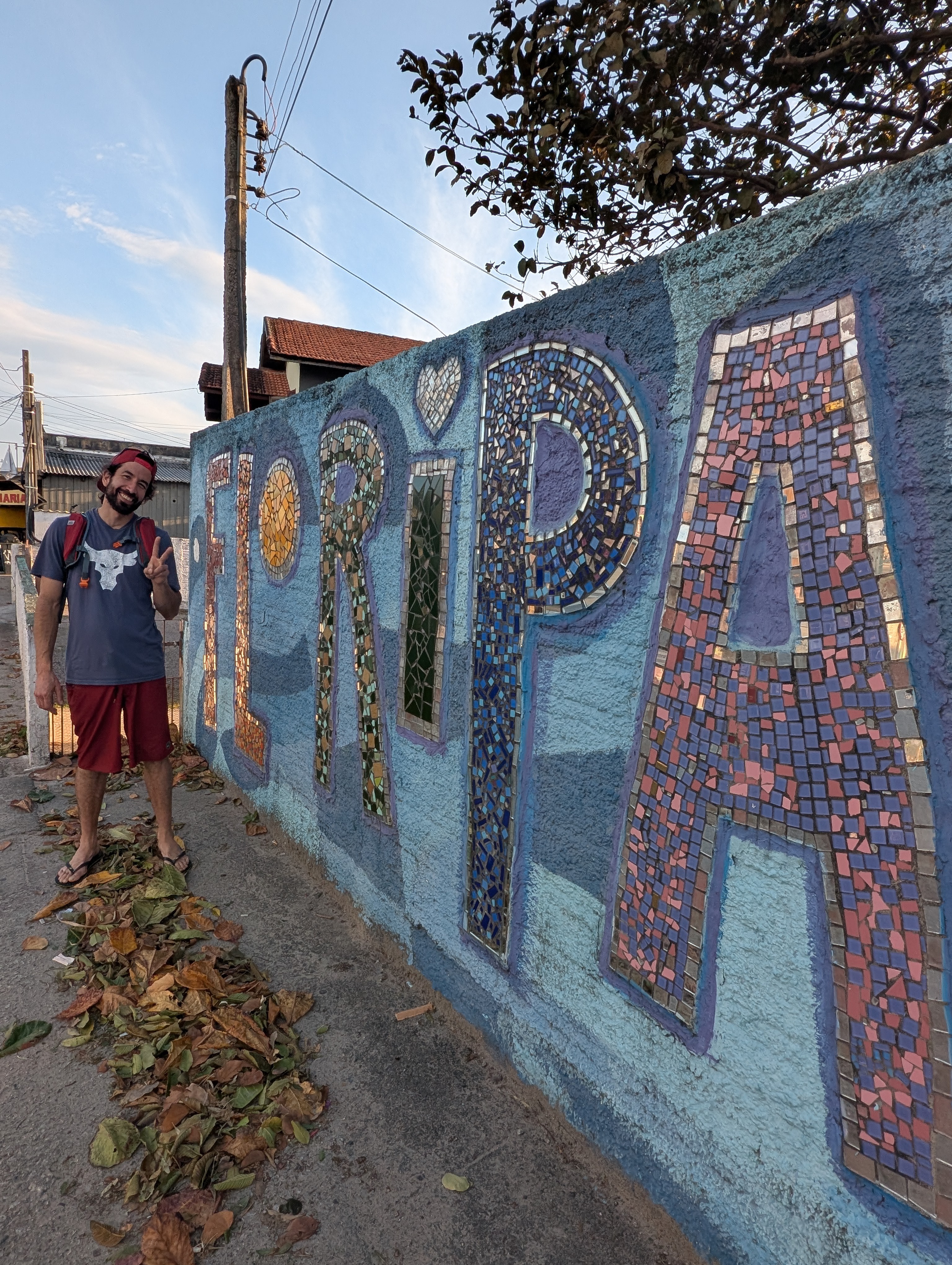 Jose Manuel flashing a peace sign beside a mosaic wall spelling Floripa