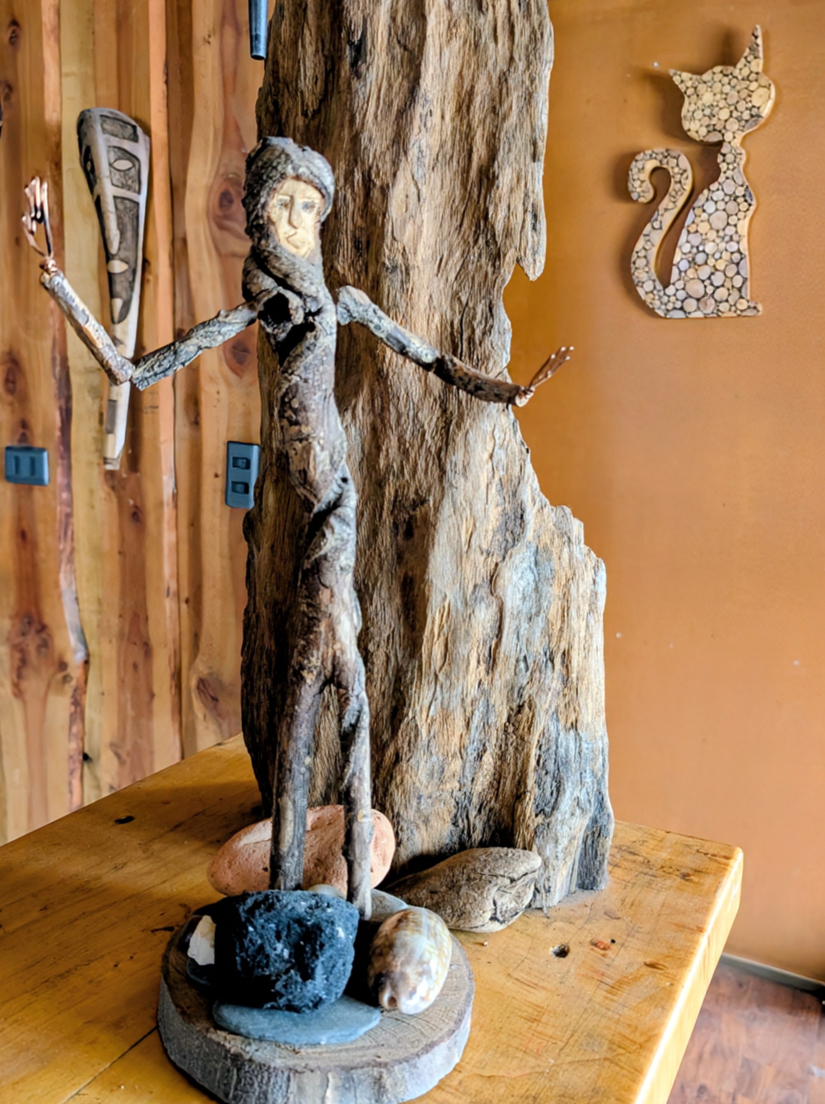 A driftwood sculpture of a figure with outstretched arms standing on stones and shells
